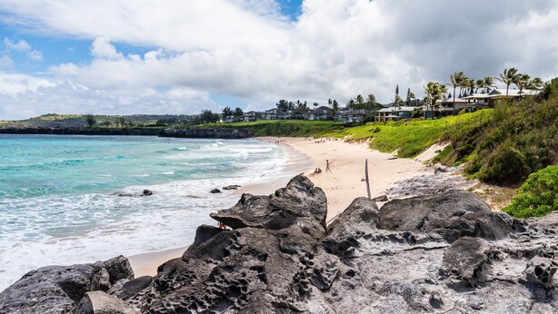 Una playa costera con primer plano rocoso, aguas turquesas transparentes y una zona residencial distante con casas y palmeras.