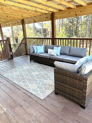A covered outdoor seating area with a wooden deck, a woven sofa with cushions, and a patterned rug. - Coastal Escape Includes Golf Cart, Bikes and Multiple Covered Outdoor Areas (Dauphin Island)
