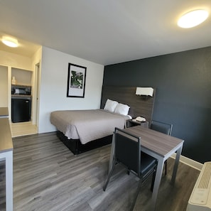A hotel room with a bed, bedside table, chair, desk, and a small kitchenette.