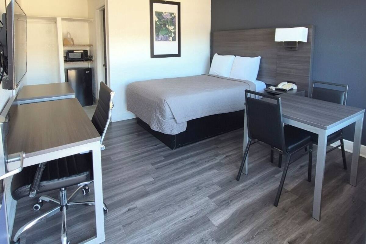 Habitación de hotel pequeña con cama, escritorio y una silla. Cuenta con una kitchenette con microondas y refrigerador.