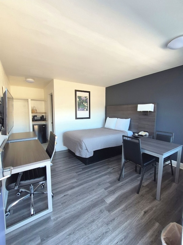 A compact hotel room with a bed, a desk, and a chair. There is a small kitchenette with a microwave and a refrigerator.
