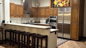 A modern kitchen with wooden cabinets, a stainless steel refrigerator, a central island with bar stools, and recessed ceiling lighting. - Mountain Side luxury Condo (Ketchum)