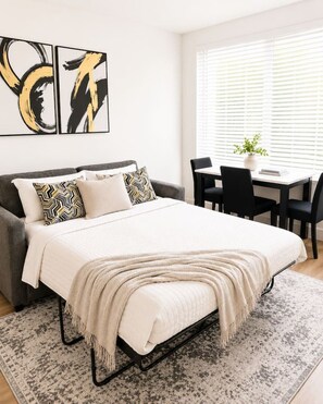 A modern bedroom with a bed, a gray sofa, a dining table with chairs, and abstract wall art. - Downtown Escape by Bathhouse Row & Convention Cntr (Hot Springs)