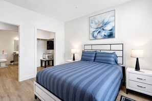 Ein Schlafzimmer mit einem Bett, Nachttischen, einer Lampe, einem Gemälde und einem durch eine offene Tür einsehbaren Badezimmer.