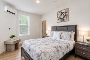 A bedroom with a bed, bedside tables, a nightstand, a wall-mounted air conditioner, a window with blinds, and a painting on the wall. - Modern City Retreat- Walk to Convention Center (Hot Springs)