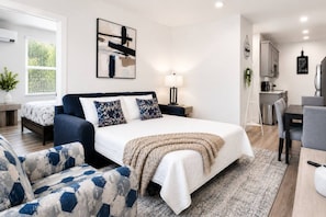 A modern bedroom with a large bed, a bedside table, a lamp, and a view of the kitchen area.