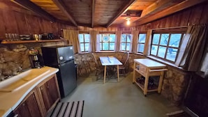 A rustic kitchen with wooden walls, a stone accent wall, a refrigerator, a sink, a wooden table, and a window with curtains.