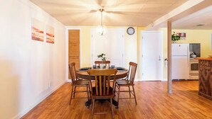 Uma sala de jantar com mesa redonda de madeira e cadeiras, um lustre e uma cozinha ao fundo.