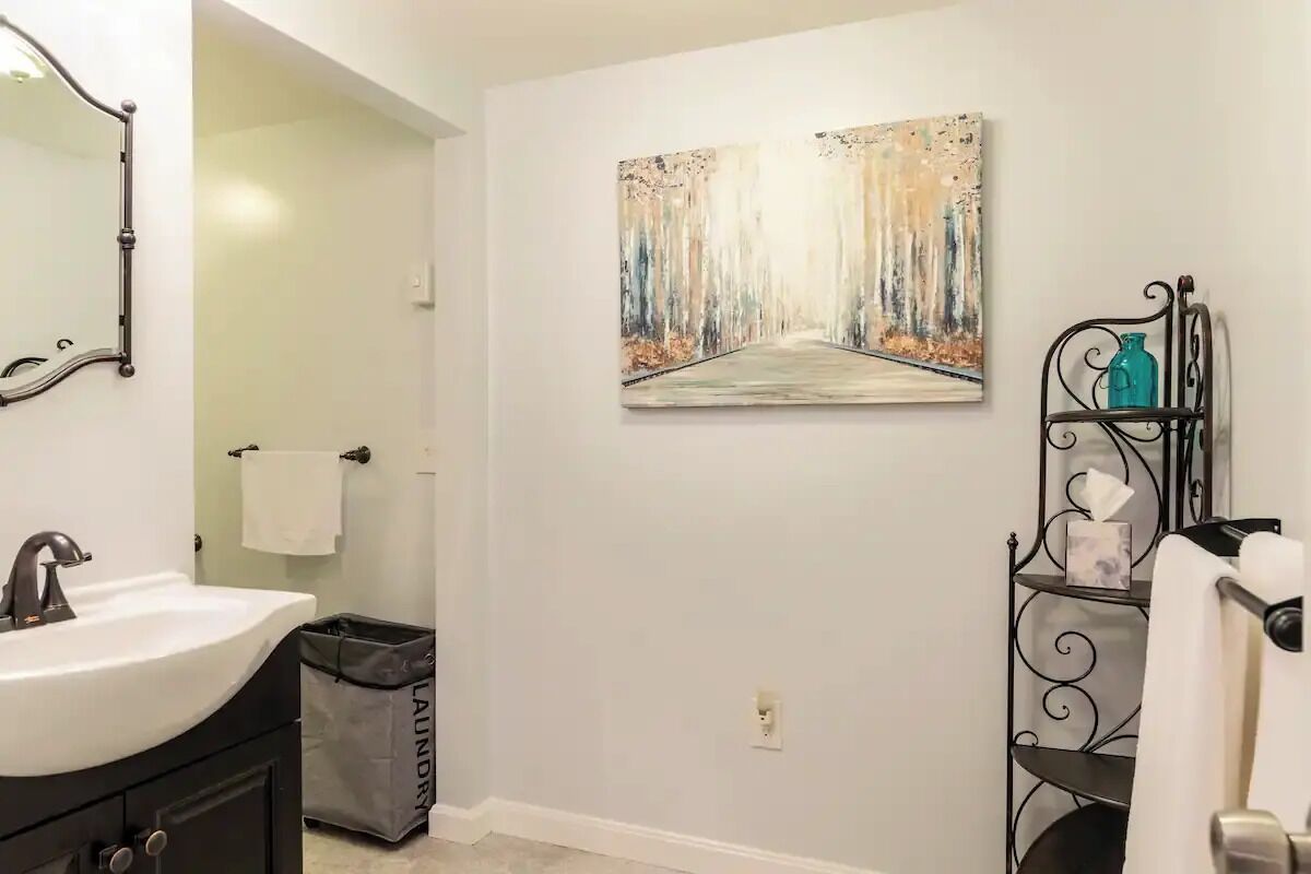 A bathroom with a white sink, a mirror, a painting of a forest path, a black metal shelf with toiletries, and a trash bin labeled "laundry".