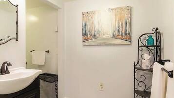 A bathroom with a white sink, a mirror, a painting of a forest path, a black metal shelf with toiletries, and a trash bin labeled "laundry".