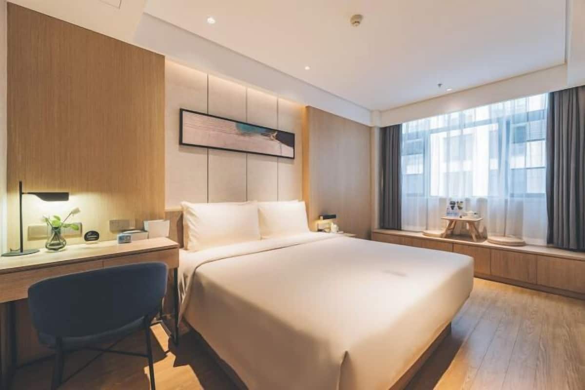 Quarto de hotel moderno com uma cama grande, uma escrivaninha com cadeira, mesa de cabeceira e uma janela ampla com cortinas.