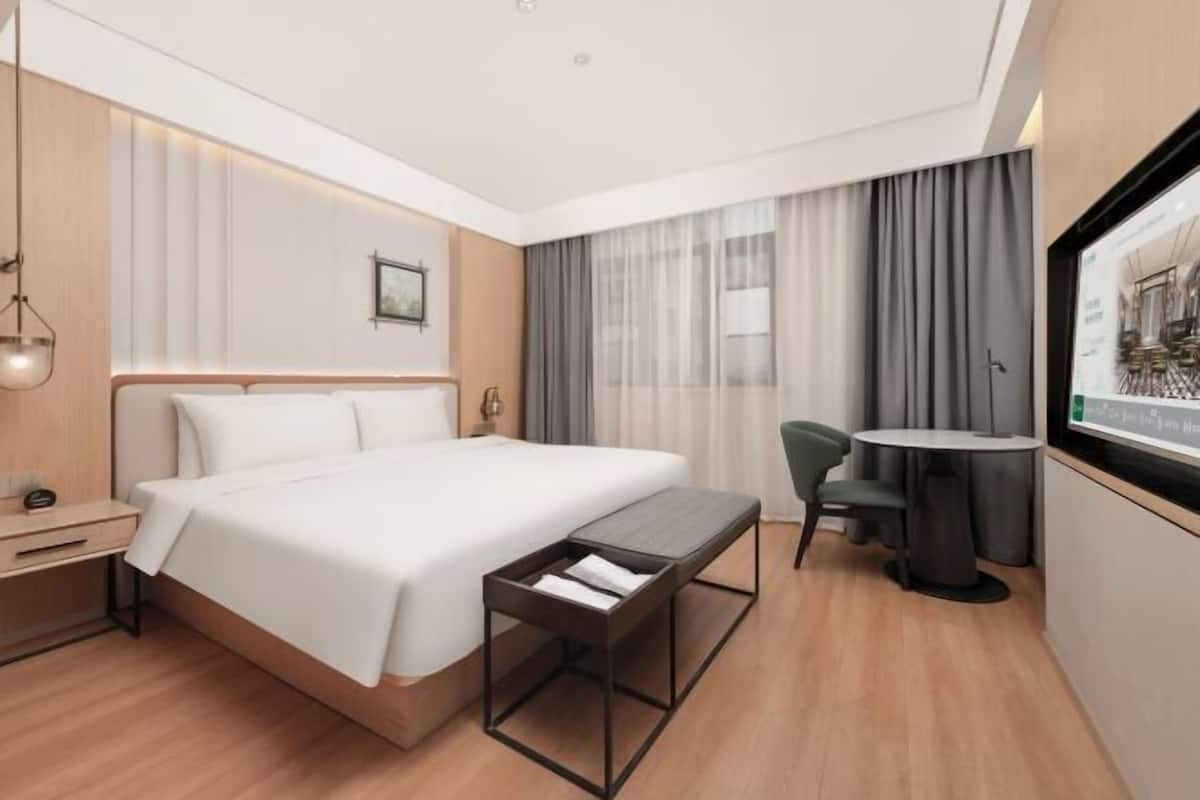 Quarto de hotel moderno com uma cama grande, mesa de cabeceira e pequena escrivaninha com uma cadeira.