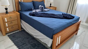 Una cama bien hecha con colcha y almohadas azules, mesitas de noche de madera con lámparas, una alfombra oscura y un suelo de mármol.