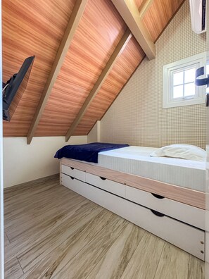 Quarto com teto inclinado, cama com manta azul, janela e ar condicionado fixado na parede.