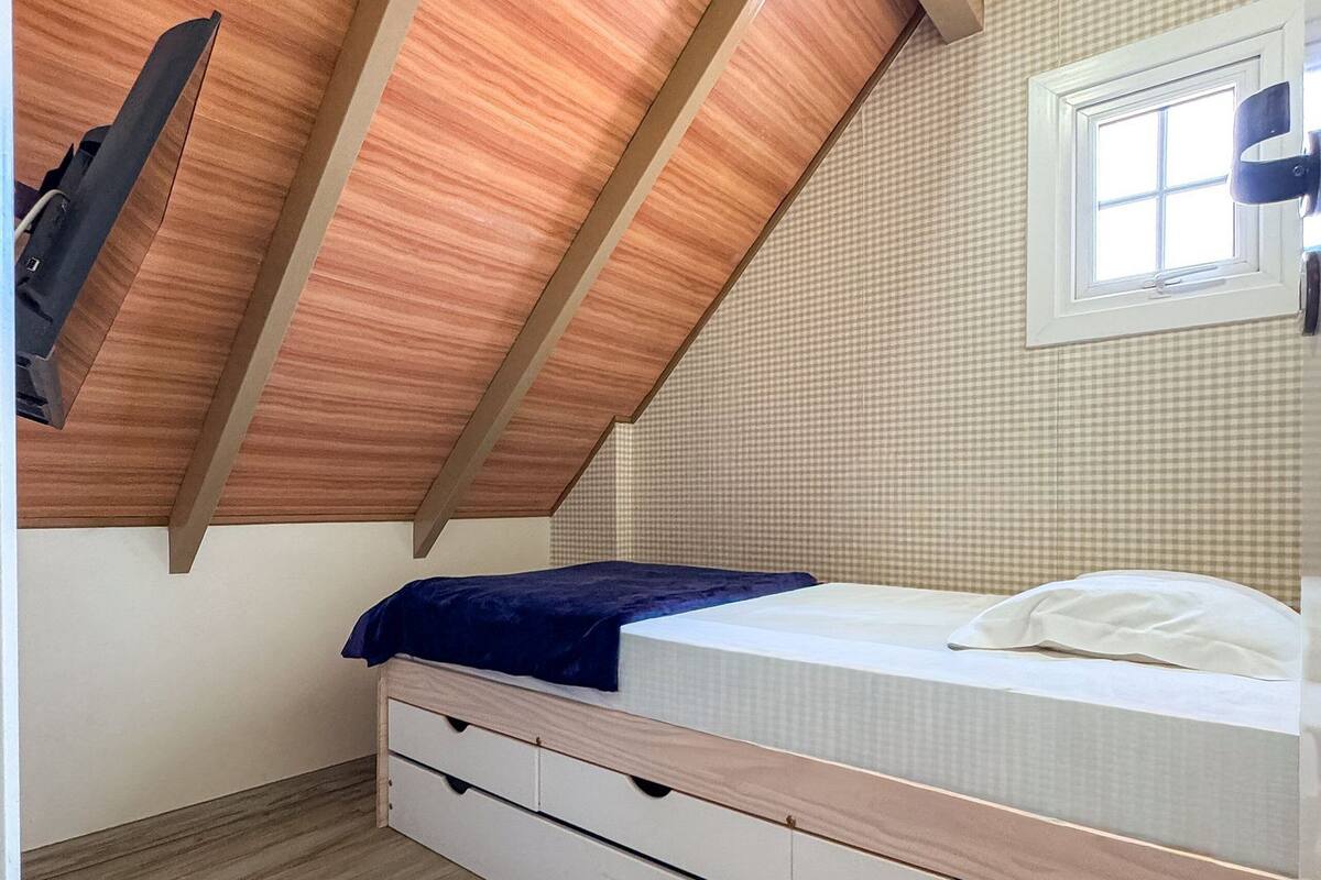 Un dormitorio con techo inclinado, una cama con manta azul, una ventana y un equipo de aire acondicionado instalado en la pared.