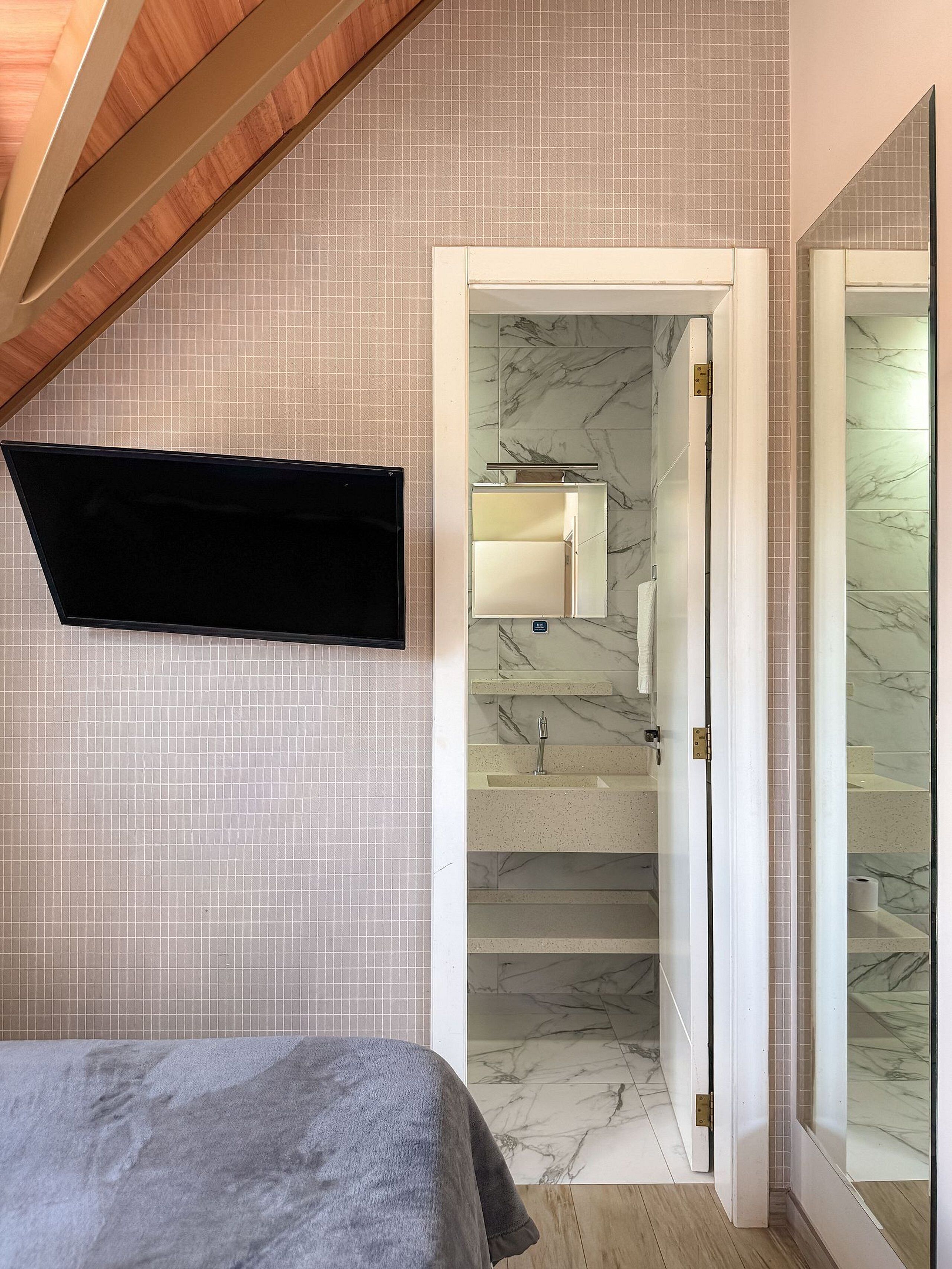 Un dormitorio con cama, televisor montado en la pared, baño con piso de mármol y una escalera con espejo.