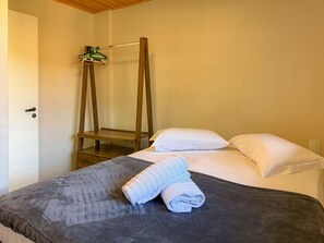 Uma cama de solteiro com um espalhador cinzento, duas almofadas brancas e uma toalha branca enrolada. Uma prateleira de madeira com um objeto verde está fixada na parede acima da cama.