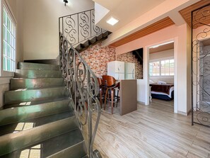 Una escalera con barandillas de metal, una zona de cocina con armarios blancos y un dormitorio con cama y ventana.