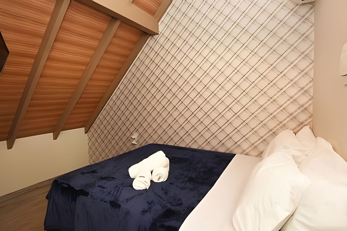A hotel room with a bed, a window with blinds, and a wooden ceiling.