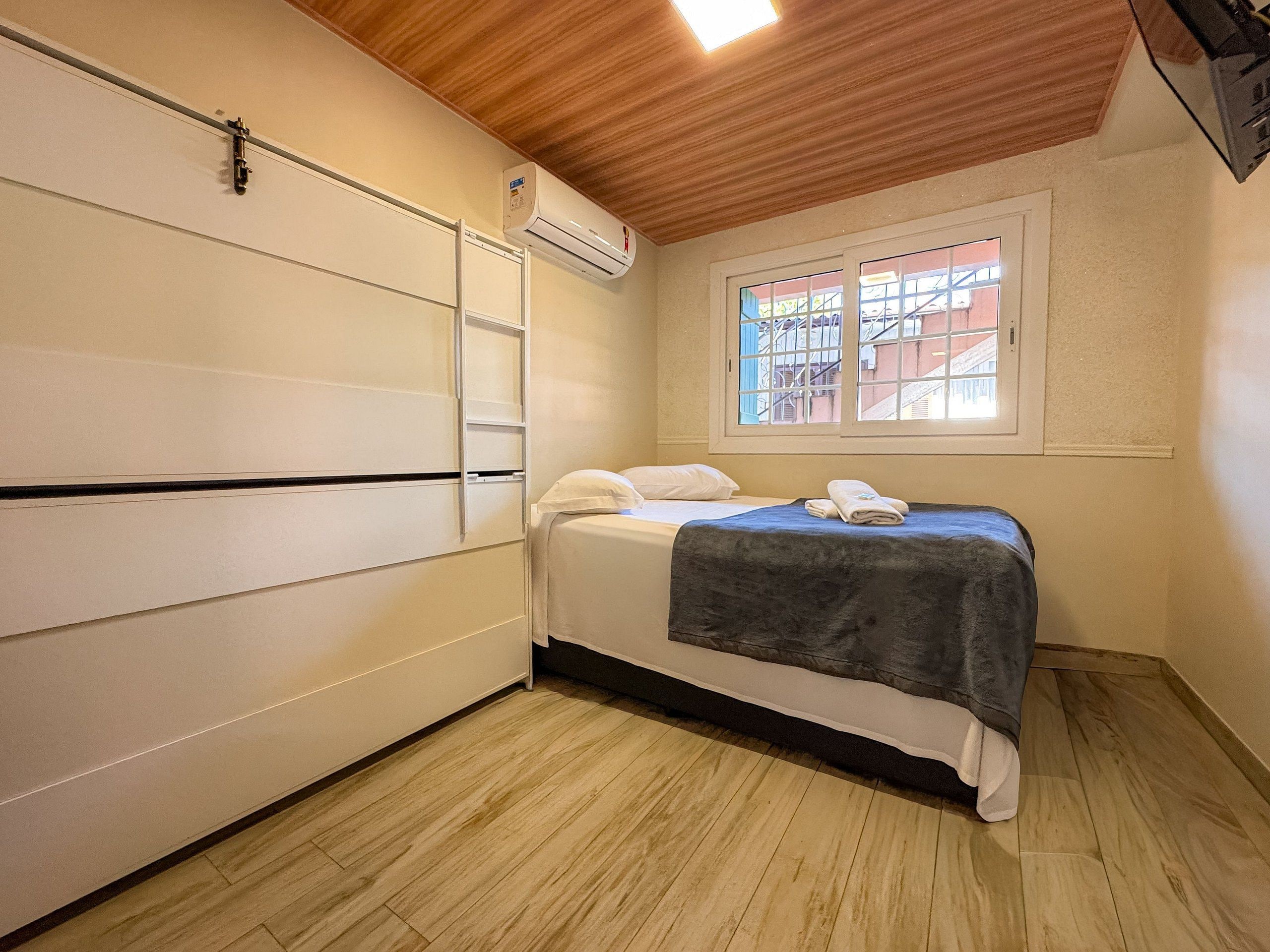 A bedroom with a bed, a window with a view, an air conditioning unit, and a wooden ceiling.