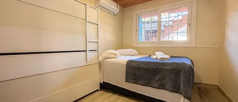 A bedroom with a bed, a window with a view, an air conditioning unit, and a wooden ceiling.