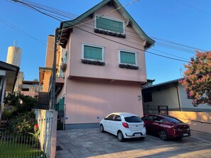 Un bâtiment à deux étages avec une façade rose, deux fenêtres dotées de volets verts et une voiture blanche garée devant.