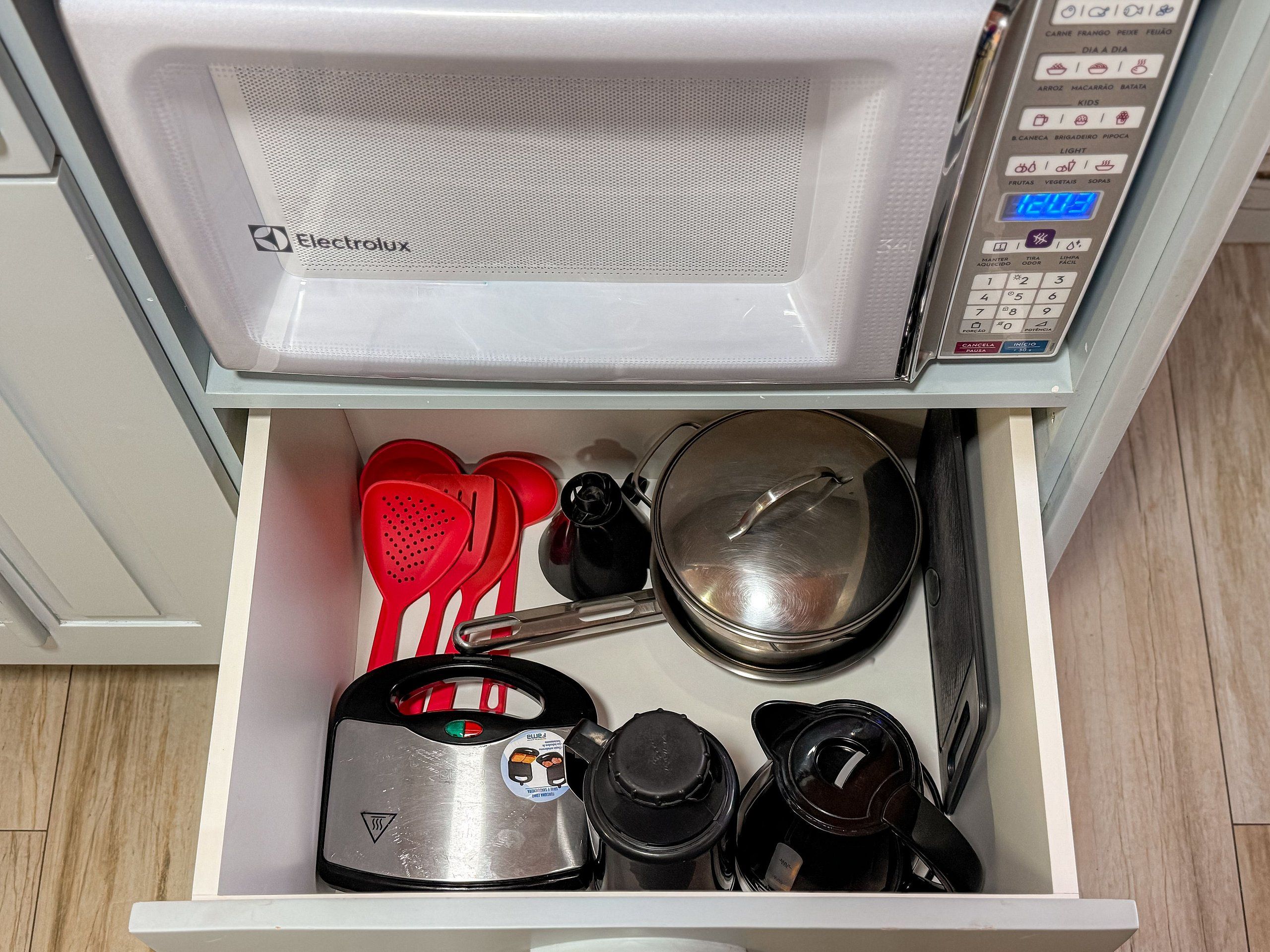 A microwave with a digital display, a kettle, a pan, and red spatulas in a drawer.