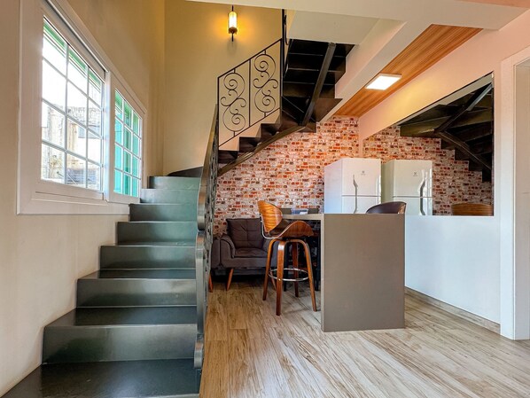 A staircase leading to a kitchen with a brick wall, a refrigerator, and a dining area with a chair.