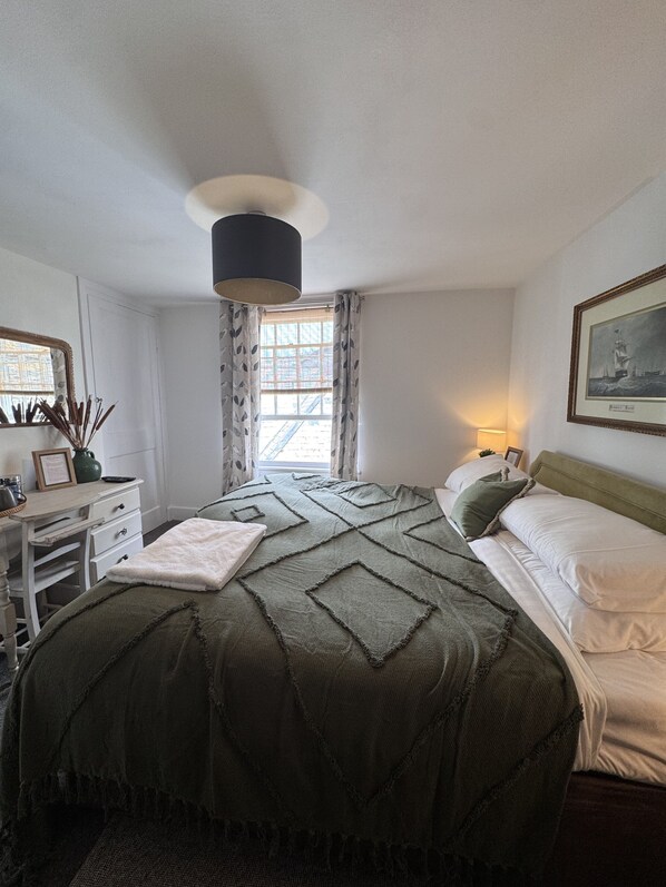 A bedroom with a bed, a desk, a lamp, a window with curtains, and a framed picture on the wall. - The Ship Inn (Mevagissey)