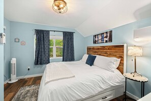 Een slaapkamer met een bed, een bedlampje, een raam met gordijnen, een wandheater en een schilderij.