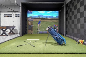 Configuración de simulación de golf virtual con zona de práctica cercada por una red, un bolso de golf y una pantalla mostrando un campo de golf.