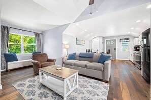 Een moderne woonkamer met een bank, fauteuil, salontafel en uitzicht op het buitenleven door een raam.