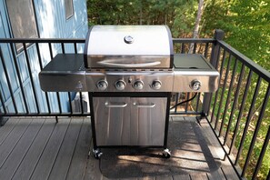 Un barbecue en acier inoxydable doté de quatre brûleurs et d’une étagère latérale, installé sur une terrasse en bois avec une rambarde.