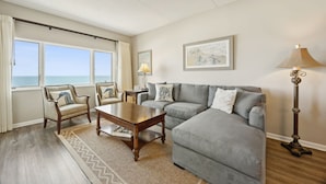 Ein Wohnzimmer mit grauem Sofa, einem Holztisch, zwei Sesseln, einer Stehlampe und Meerblick durch große Fenster.