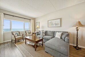 Ein Wohnzimmer mit grauem Sofa, einem Holztisch, zwei Sesseln, einer Stehlampe und Meerblick durch große Fenster.