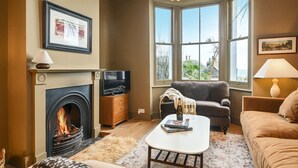 A cozy living room with a fireplace, a sofa, a coffee table, and a view through large windows.