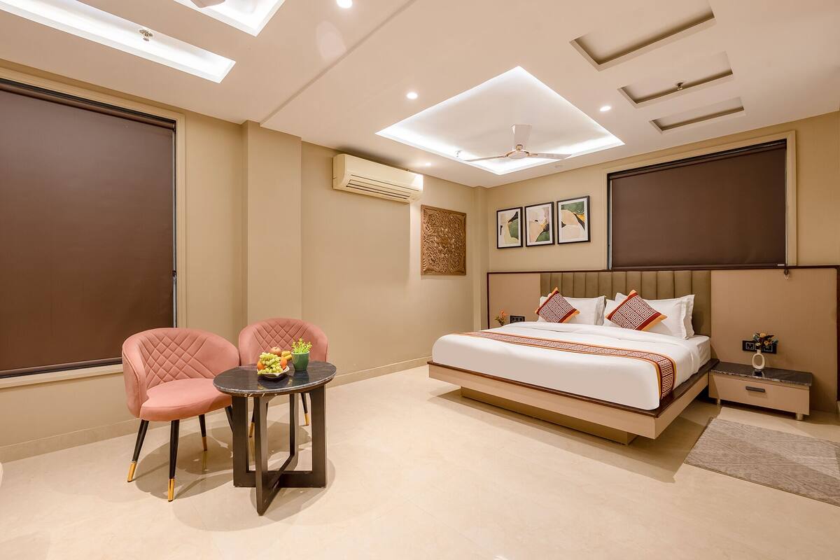 A hotel room with a bed, two chairs, a small table, a ceiling fan, recessed lighting, artwork, and an air conditioning unit.