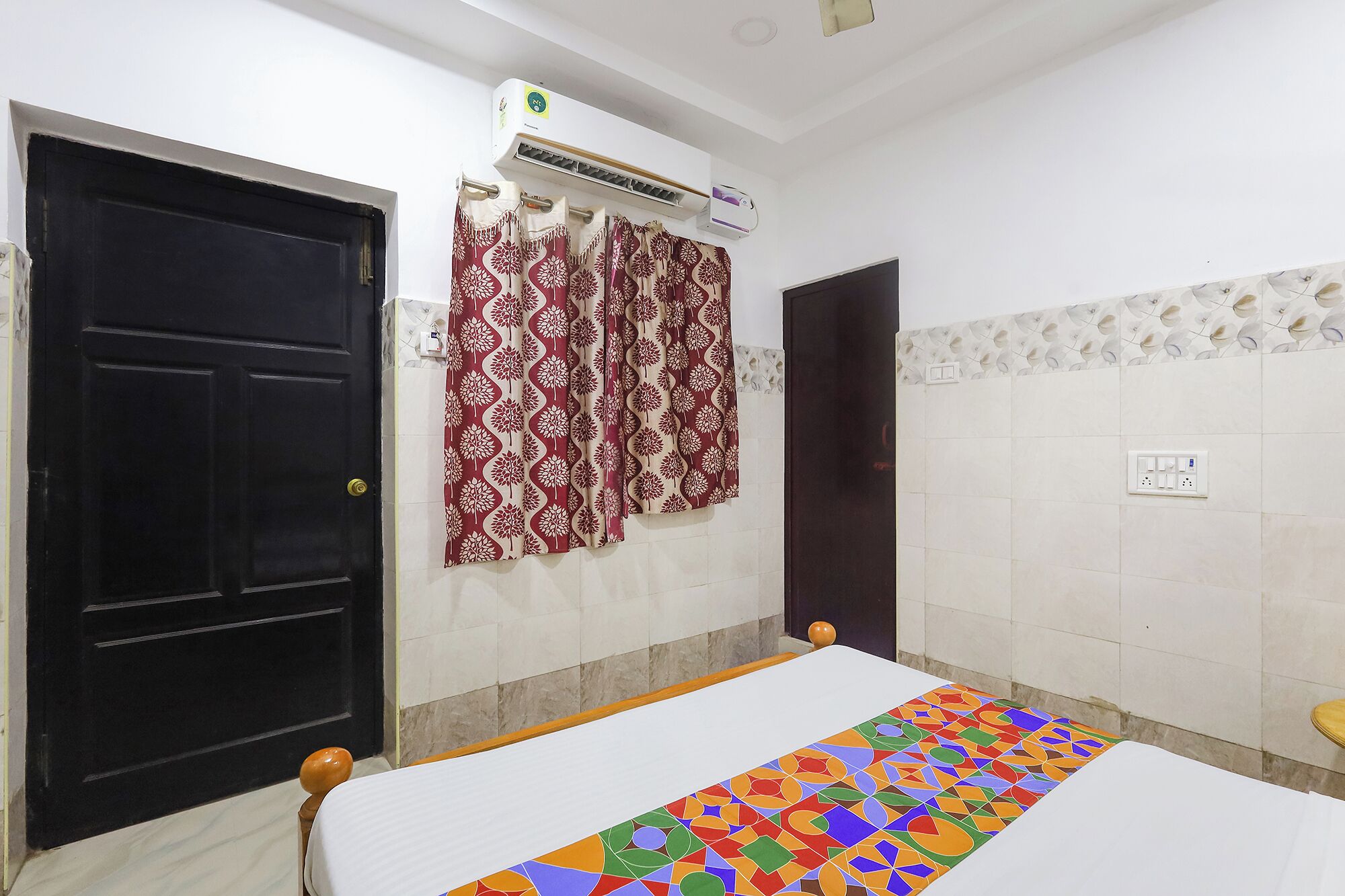A room with a bed, an air conditioning unit, patterned curtains, and a tiled wall.