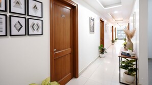 A hallway with a wooden door labeled 102, potted plants, framed artwork on the walls, and a small table with books and a vase. - FabHotel Expressway 135 - Near By KJR Hospital (Noida)