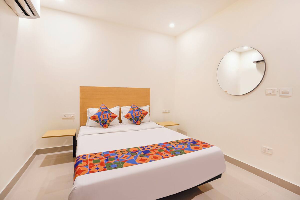 Een moderne hotelkamer met een houten hoofdeinde, een groot bed met een kleurrijke, geometrische spreideken, twee decoratieve kussens, een ronde spiegel en een klein houten nachtkastje.
