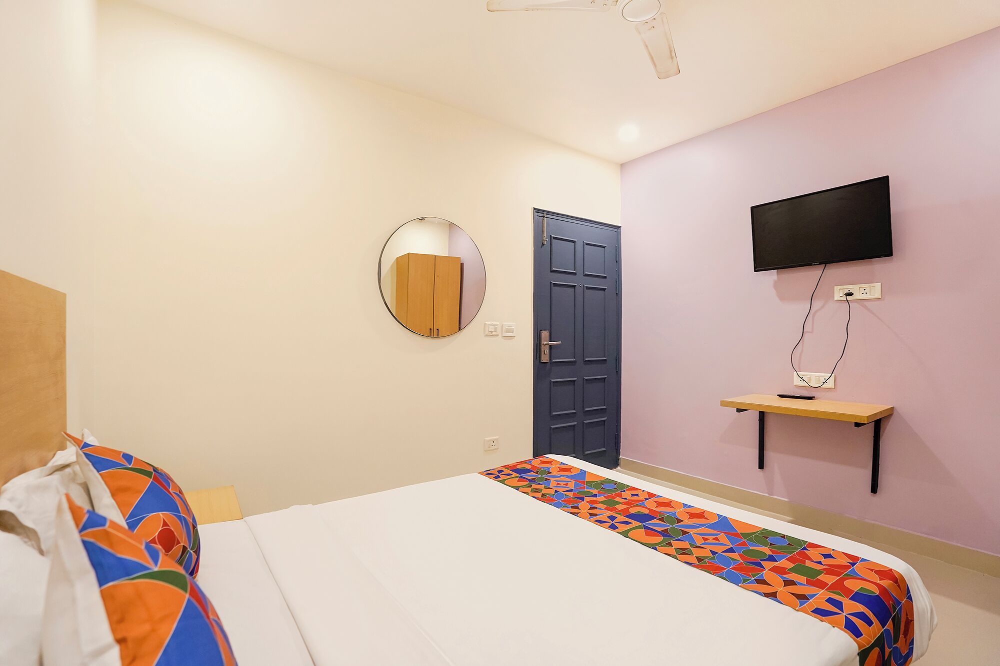 Habitación de hotel con cama, un espejo redondo, una puerta, un televisor montado en la pared y una mesita.
