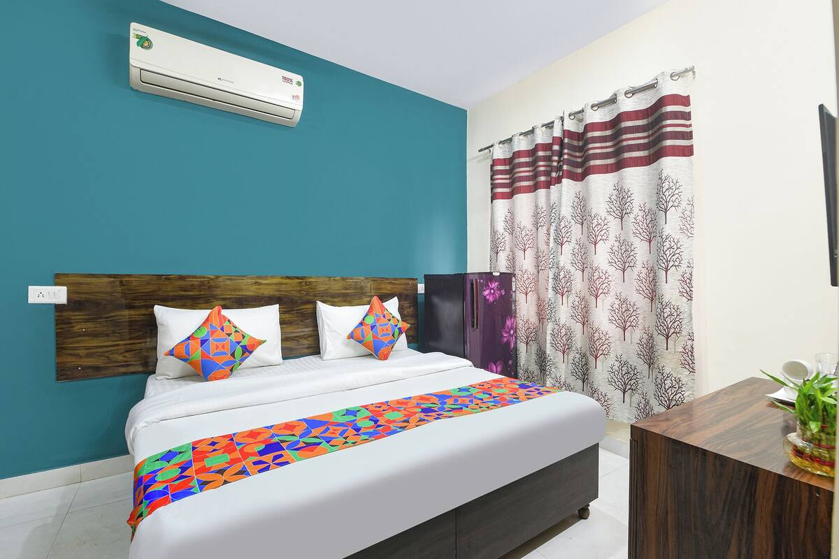 A bedroom with a bed, a wooden headboard, a wall-mounted air conditioner, a patterned curtain, and a small wooden side table with a plant.