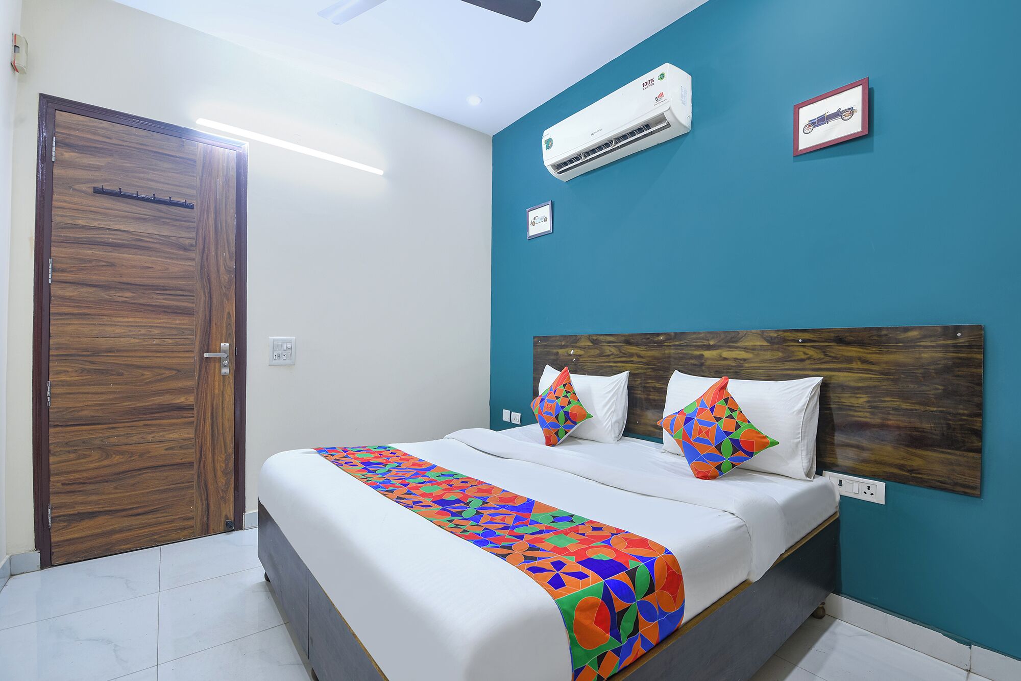 A hotel room with a wooden door, a bed with white bedding and colorful pillows, an air conditioning unit, and a teal accent wall.