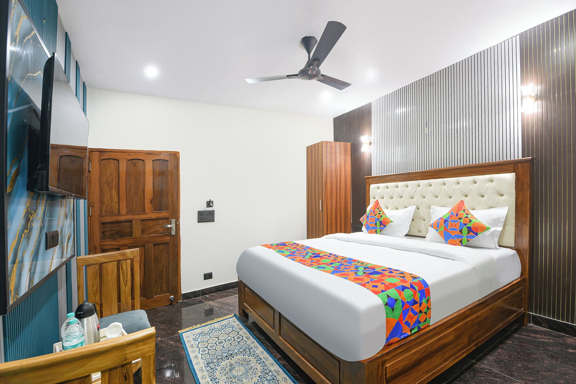 A bedroom with a bed, a ceiling fan, wooden wardrobe, and a small table with a water bottle.