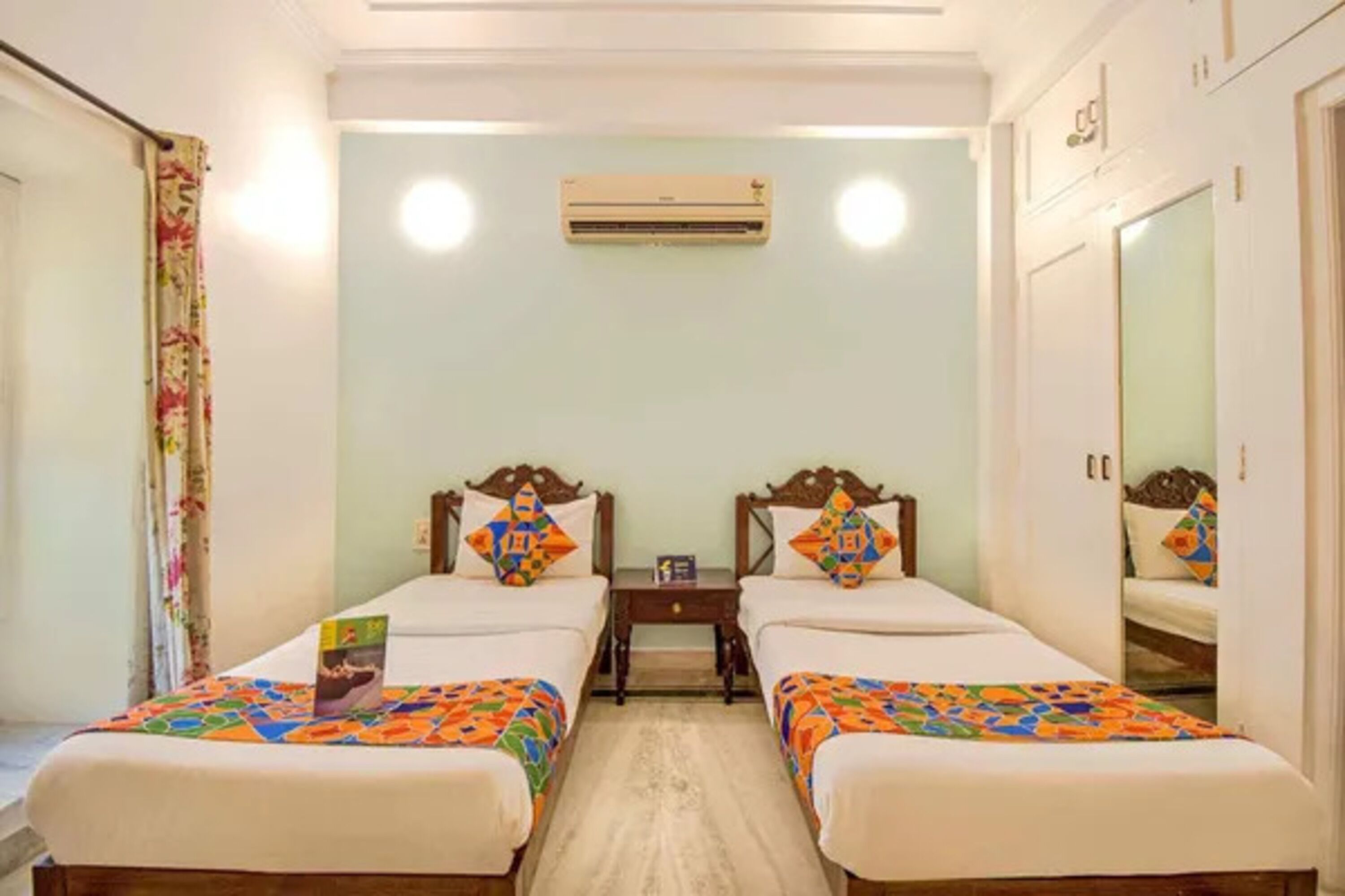 A hotel room with two single beds, a small bedside table, an air conditioning unit, and a mirror on the wall.