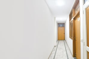 Un pasillo estrecho con una puerta de madera al final, paredes blancas y suelo de baldosas.