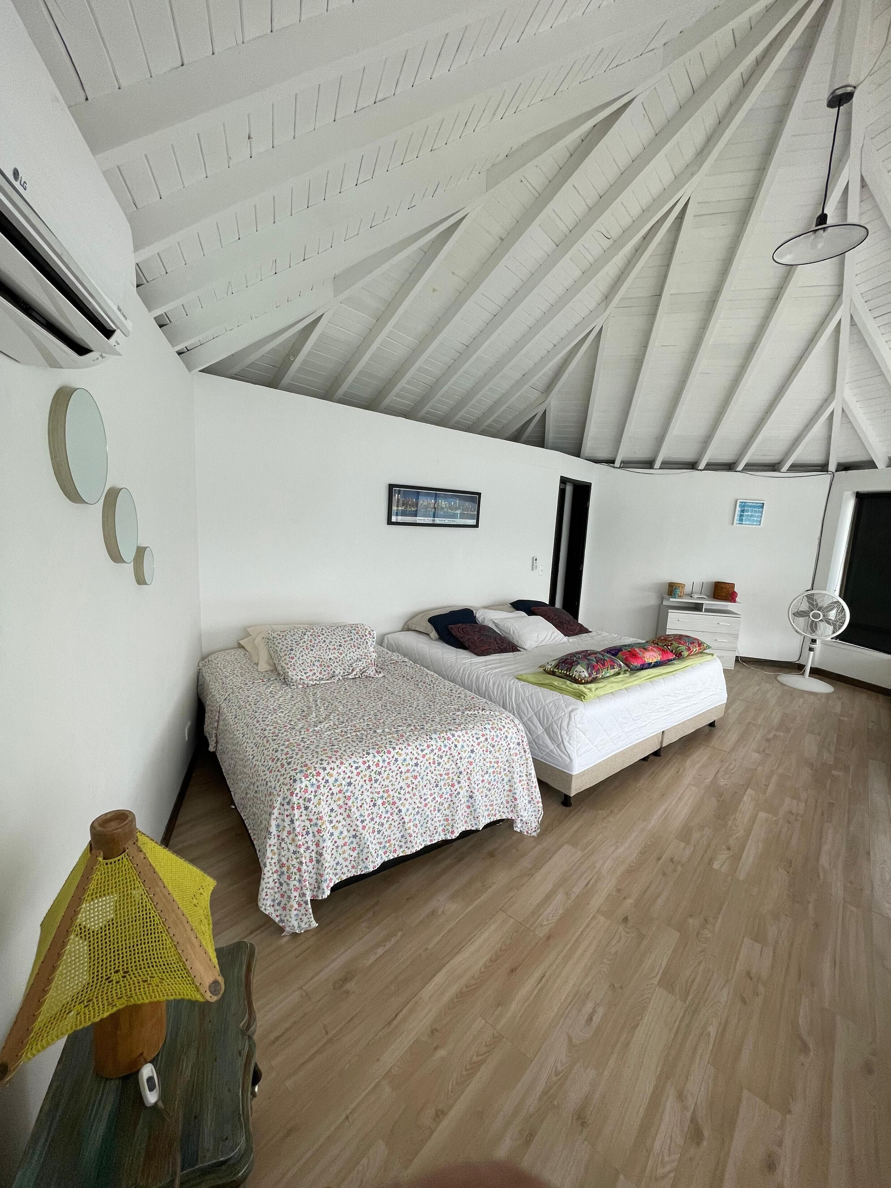 Un dormitorio con techo inclinado, cama con colcha estampada, mesita de noche con lámpara y aire acondicionado de pared.