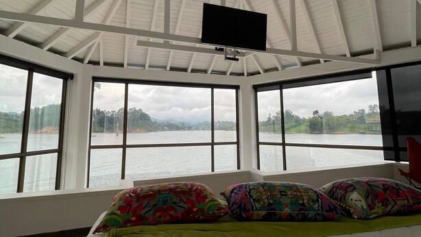 A room with a bed, a television mounted on the ceiling, and large windows offering a view of a body of water and hills. - Finca La Cruz (El Penol)