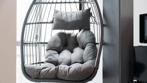 A grey upholstered hanging chair with a cushion, positioned against a wall with a window and a flat-screen TV.