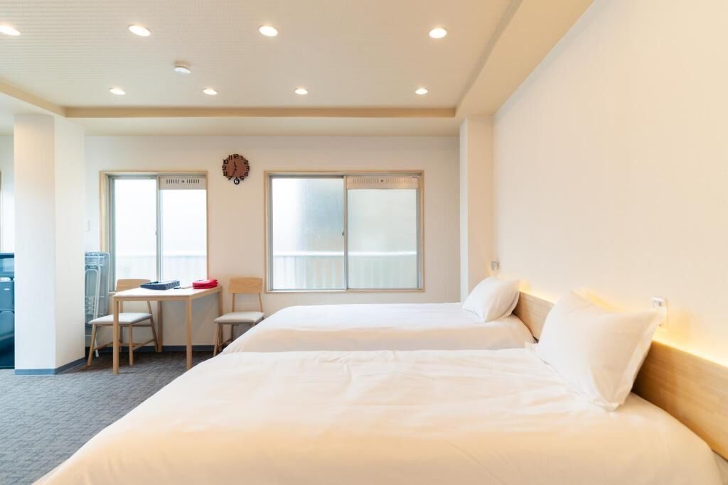 A hotel room with two beds, a small table with chairs, a clock on the wall, and large windows.